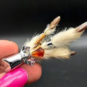 Peut inclure: Épingle de kilt argentée avec une tête décorative ornée d'un motif animal, de perles ambrées et de touffes de fourrure et de plumes claires. L'épingle est tenue par une personne.