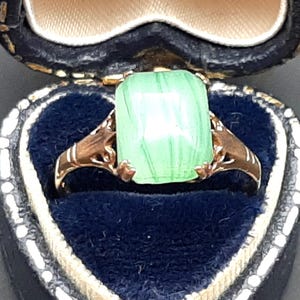 May include: A gold ring featuring a rectangular, pale green gemstone. The ring is presented in a blue velvet jewellery box. The gemstone has a striped pattern. The ring has an ornate, vintage design.