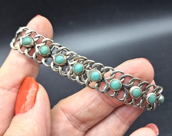 Op de afbeelding: Een zilverkleurige metalen schakelarmband met een herhalend opengewerkt patroon. De armband is bezet met een reeks kleine, ronde, gladde, lichtturquoise cabochonstenen die in een enkele lijn in het midden van de metalen schakels zijn geplaatst.