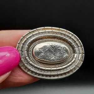 May include: An oval, antique silver brooch with a floral design in the center. The brooch has a detailed, layered border with a textured pattern. The piece is held by a hand with pink nail polish against a black background.