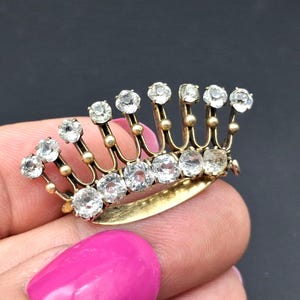 Op de afbeelding: Een goudkleurige metalen broche in de vorm van een kroon, met een gebogen basis en verticale pinnen bekroond met heldere, ronde, gefacetteerde stenen. Het ontwerp bevat kleine gouden bolletjes aan de basis van elke pin voor een gestructureerde uitstraling.