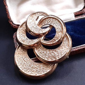 Può includere: Spilla in metallo dorato con un design astratto a spirale. La spilla è composta da più cerchi intrecciati con una superficie testurizzata.