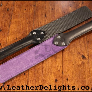 May include: Two leather paddles with different surfaces. One paddle has a black leather handle and a purple suede surface. The other paddle has a black leather handle and a black textured surface. Both paddles have silver metal accents.
