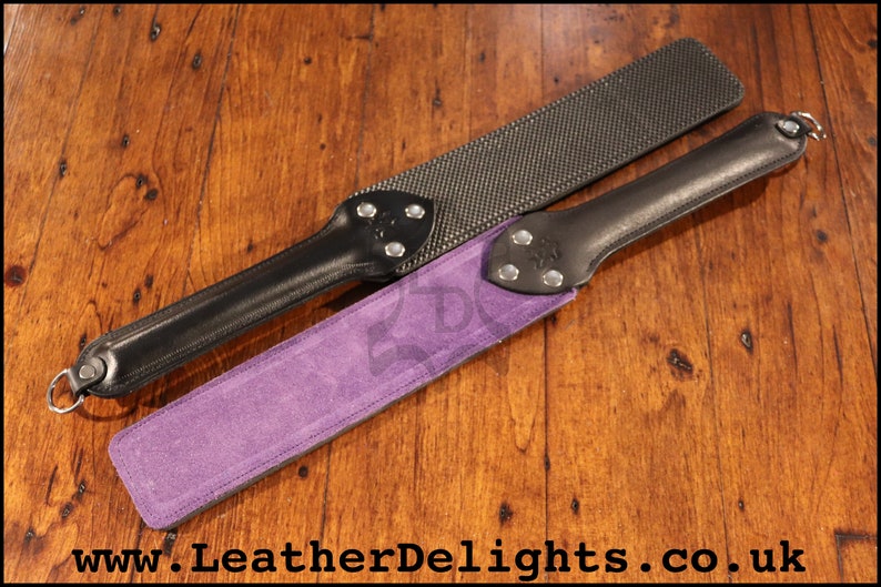 May include: Two leather paddles with different surfaces. One paddle has a black leather handle and a purple suede surface. The other paddle has a black leather handle and a black textured surface. Both paddles have silver metal accents.