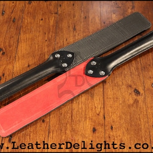 May include: Two black leather paddles with different striking surfaces. One paddle has a black textured surface and the other has a red surface. Both paddles have a black leather handle with a metal ring at the end.