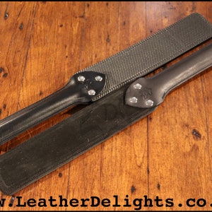 May include: Two black leather paddles with metal accents. The paddle on top has a textured surface. The paddle on the bottom has a smooth surface. The paddles are on a wooden surface.