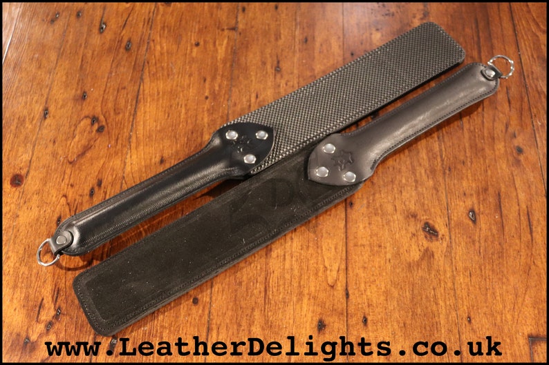 May include: Two black leather paddles with metal accents. The paddle on top has a textured surface. The paddle on the bottom has a smooth surface. The paddles are on a wooden surface.