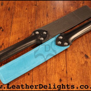 May include: Two leather paddles with different surfaces. One paddle has a black leather handle and a blue suede surface. The other paddle has a black leather handle and a textured black surface. Both paddles have silver metal accents.