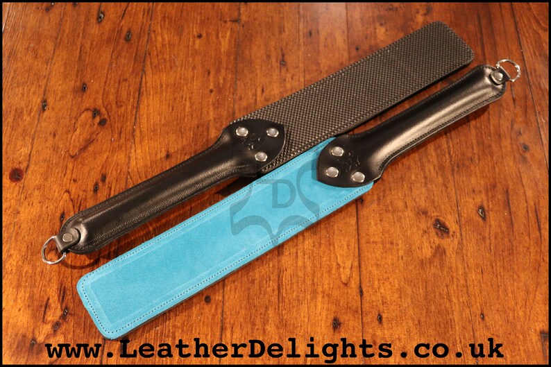 May include: Two leather paddles with different surfaces. One paddle has a black leather handle and a blue suede surface. The other paddle has a black leather handle and a textured black surface. Both paddles have silver metal accents.