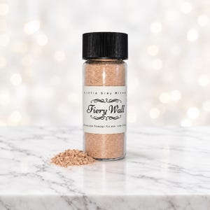 Peut inclure: Un flacon en verre transparent avec un couvercle noir à vis strié contenant une poudre brun clair. L'étiquette blanche porte le texte Little Grey Witch, Fiery Wall et Conjure Powder for ext. Use only. Un petit tas de poudre est posé à côté.
