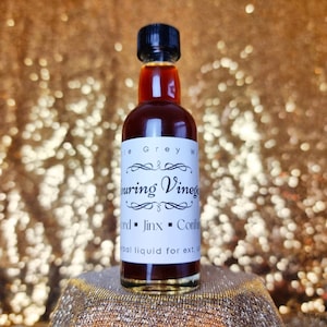 Op de afbeelding: Een klein glazen flesje gevuld met een donkerbruine vloeistof. Het flesje heeft een wit etiket met zwarte tekst die "White Grey Wine Souring Vinegar - Ward - Jinx - Conjure - Herbal liquid for ext. use" luidt.