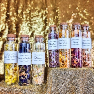 Puede incluir: Seis pequeñas botellas de vidrio llenas de flores y hierbas secas. Las botellas están etiquetadas con los nombres de las flores y hierbas, incluyendo Siempreviva, Malva azul, Altea, Brezo, Calendula, Rosa y Jazmín.