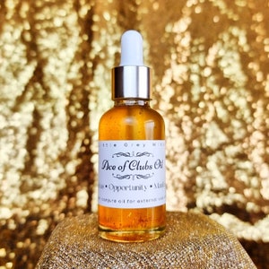 Può includere: Una bottiglia di vetro trasparente con un tappo argentato e un contagocce riempita di un liquido dorato. L'etichetta della bottiglia riporta "Ace of Clubs Oil" e elenca "Fortuna, Opportunità, Manifestazione" come benefici. La bottiglia è un olio di magia per uso esterno.