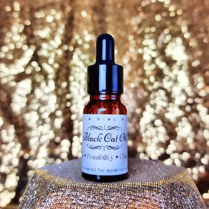 May include: A small brown glass bottle with a black dropper top. The bottle is labeled "Black Cat Oil" and says "Possibility - Change" in smaller text. The bottle is sitting on a gold sparkly surface.