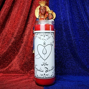 Puede incluir: Vela roja con una etiqueta blanca que presenta un corazón con una daga atravesándolo y el texto "Little Gypsy Witch" y "Ezjulie Dantor".