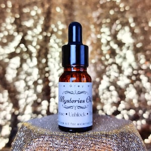 Puede incluir: Un pequeño frasco de vidrio marrón con un gotero negro. El frasco está etiquetado con "Mysteries Oil" y tiene el texto "Unblock" en la etiqueta. El frasco está sobre una superficie dorada.