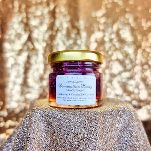 Puede incluir: Un pequeño frasco de vidrio con tapa dorada lleno de purpurina morada y miel. La etiqueta dice "Little Grey Witch Domination Honey Command Compell Control Honey blended with oils and herbs for external use only."