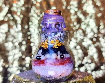 Puede incluir: Una botella de vidrio con un sello de cera púrpura, llena de varios cristales, cuentas y flores secas. La botella está sobre una superficie dorada.