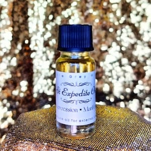 Può includere: Una piccola bottiglia di vetro con tappo nero, riempita con un liquido color oro. L'etichetta recita "St Expedite Oil" con testo aggiuntivo. La bottiglia è esposta su una superficie dorata con paillettes.