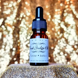 Può includere: Un piccolo flacone di vetro marrone con un contagocce nero. L'etichetta recita "Just Fudge Oil" con le parole "Favour - Justice" sotto. Il flacone è appoggiato su una superficie dorata scintillante.