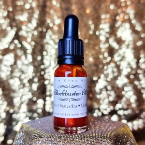 May include: A small, clear glass bottle with a black dropper top. The bottle is filled with a red liquid and has a white label that reads "Blockbuster Oil", "Move Obstacles", "Manifest", and "Pure oil for external use".