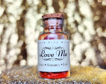 Puede incluir: Una pequeña botella de vidrio con tapón de corcho, llena de contenido rojo. La etiqueta dice "Love Me" con las palabras "Love, Romance, Lust" debajo. La botella está etiquetada como "Little Grey Witch".