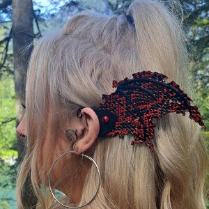 May include: A red and black leather dragon wing ear cuff with a red bead accent. The ear cuff is worn on the left ear of a person with blonde hair.
