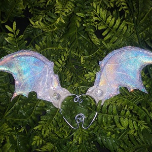 May include: A pair of iridescent silver wings with a glitter finish. The wings are attached to a wire frame and have a small crystal embellishment on each wing.