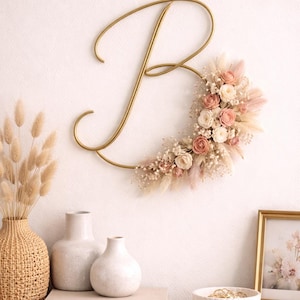 Könnte beinhalten: Goldfarbene Metall-Buchstabe "B" Wanddekoration mit einem Blumengesteck aus rosa und cremefarbenen Blumen und getrockneten Gräsern. Die Dekoration ist an einer weißen Wand befestigt. Dekorative Vasen und Bücher stehen auf einer Holzoberfläche.