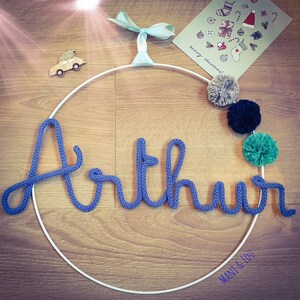 May include: A white metal hoop with the name "Arthur" spelled out in blue yarn. The hoop is decorated with pom-poms in gray, black, and green.