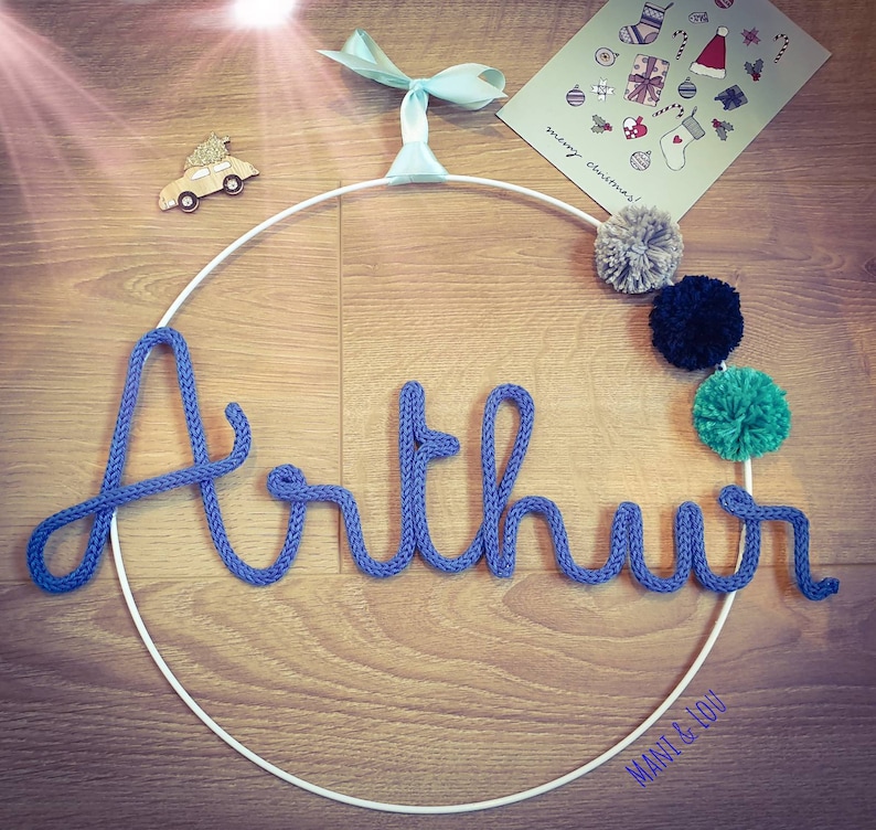 May include: A white metal hoop with the name "Arthur" spelled out in blue yarn. The hoop is decorated with pom-poms in gray, black, and green.