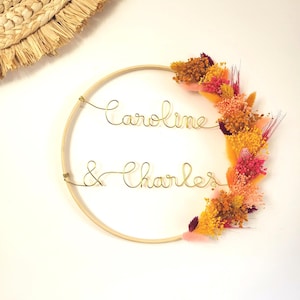 May include: A wooden hoop with the names "Caroline & Charles" written in gold wire. The hoop is decorated with dried flowers in shades of orange, pink, and yellow.