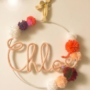 May include: A white metal hoop with the name "Chloe" spelled out in pink yarn. The hoop is decorated with pom-poms in various shades of pink, red, and white.