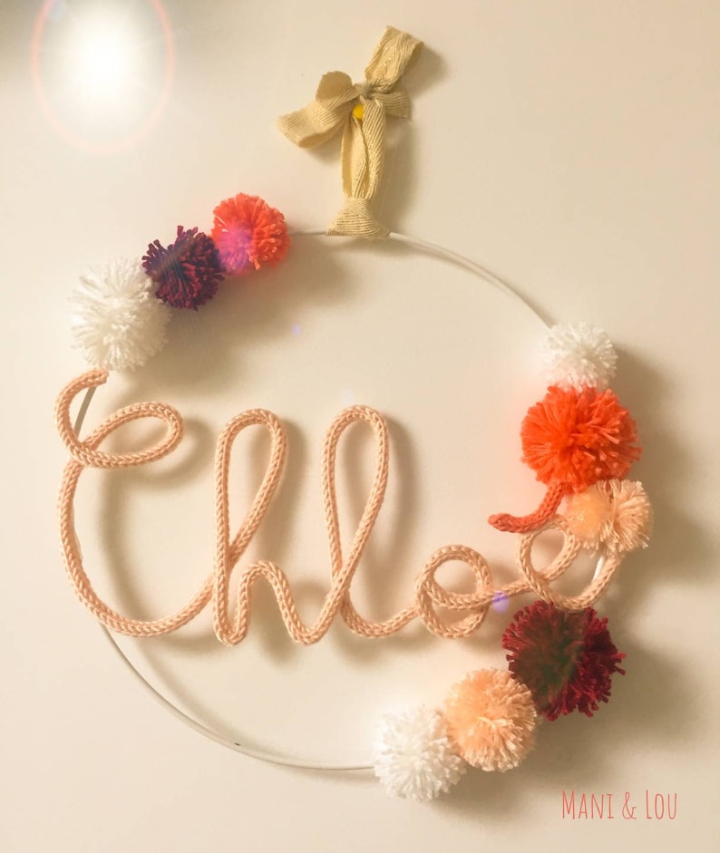 May include: A white metal hoop with the name "Chloe" spelled out in pink yarn. The hoop is decorated with pom-poms in various shades of pink, red, and white.