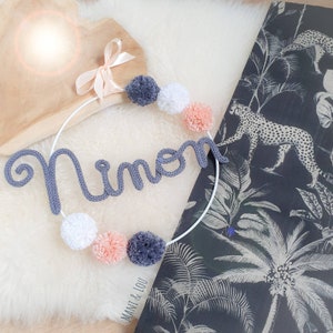 May include: A white circular hoop featuring the knitted name Ninon in grey yarn, accented with six pom-poms in shades of grey, white, and peach. A light peach ribbon is tied at the top of the hoop, which includes the text Mani & Lou printed below.