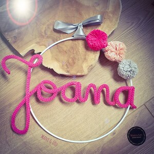 May include: Pink yarn name sign with the name "Joana" on a white metal hoop with a silver bow and pink, peach, and gray pom poms. The sign is on a wooden surface. The text "Mani & Lou" is on the hoop.