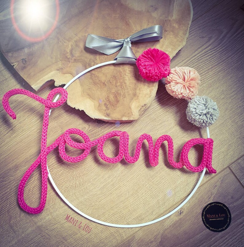 May include: Pink yarn name sign with the name "Joana" on a white metal hoop with a silver bow and pink, peach, and gray pom poms. The sign is on a wooden surface. The text "Mani & Lou" is on the hoop.