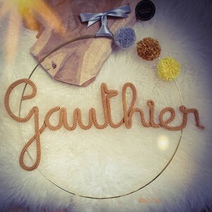 May include: A gold metal hoop with the name "Gauthier" spelled out in brown yarn. The hoop is decorated with pom-poms in shades of gray, yellow, and brown.