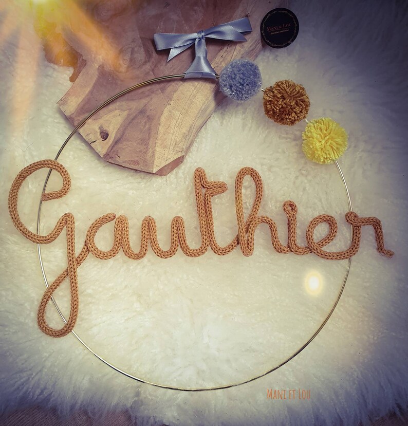 May include: A gold metal hoop with the name "Gauthier" spelled out in brown yarn. The hoop is decorated with pom-poms in shades of gray, yellow, and brown.