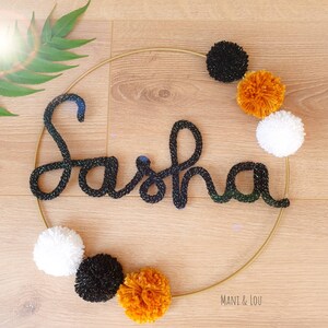 May include: A gold hoop with the name "Sasha" spelled out in black yarn. The hoop is decorated with black, white, and orange pom-poms.