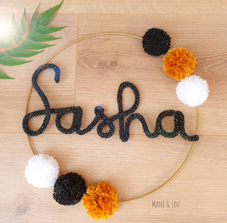 May include: A gold hoop with the name "Sasha" spelled out in black yarn. The hoop is decorated with black, white, and orange pom-poms.