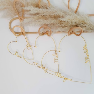 May include: Four gold wire heart-shaped hangers with the names "Maddy", "Morgan", "William", and "Chloe Love" written in the wire. The hangers are hanging on a light brown wooden rack.