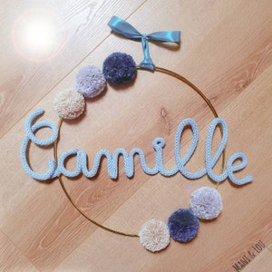 May include: A light blue knitted name sign spelling "Camille" hangs on a gold hoop with a blue ribbon. The hoop is decorated with a string of pastel pom-poms.