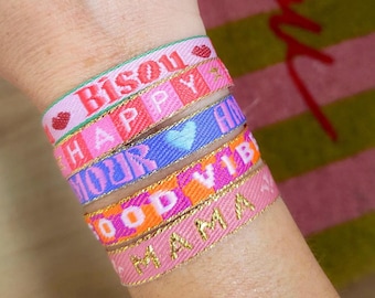 Op de afbeelding: Een stapel kleurrijke geweven armbanden met gouden randen. De armbanden tonen woorden als "Bisou", "Happy", "Your", "Good Vibes" en "Mama". De armbanden zijn roze, blauw, oranje en groen.