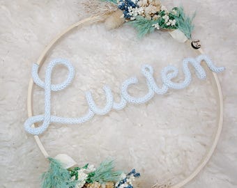 May include: A circular wooden embroidery hoop featuring the name Lucien written in white knitted yarn. The hoop is decorated with dried flowers in shades of mint green, blue, and cream. A small circular tag with the text Mani & Lou is attached.