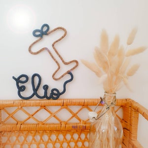 Könnte beinhalten: Ein blaues Seil-Namensschild mit der Aufschrift "Elio" und einem goldenen Seil-Flugzeug darüber. Das Schild befindet sich auf einem Weidenregal mit einer Glasflasche gefüllt mit getrocknetem Pampasgras.