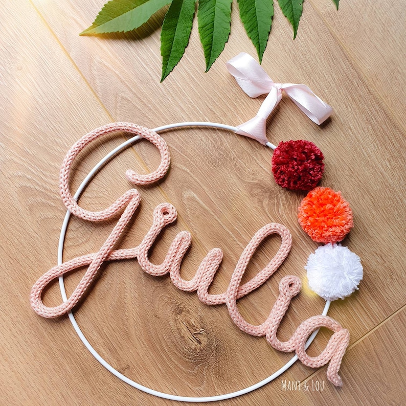 May include: A pink knitted name sign spelling "Giulia" on a white metal hoop with three pom-poms in red, orange, and white.
