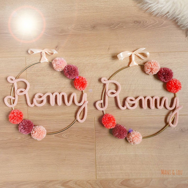 May include: Two gold hoops with the name "Rommy" spelled out in pink yarn. Each hoop is decorated with pink, red, and peach pom-poms.