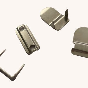 May include: A collection of silver-colored metal picture hanging hardware. The hardware includes rectangular plates with holes, curved brackets with a single nail, and L-shaped brackets with two nails. The items are scattered on a light background.