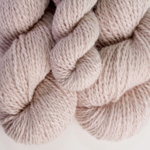 May include: Close-up of a skein of light beige yarn. The yarn is tightly wound and has a soft, textured appearance. The neutral color makes it suitable for various knitting and crochet projects.